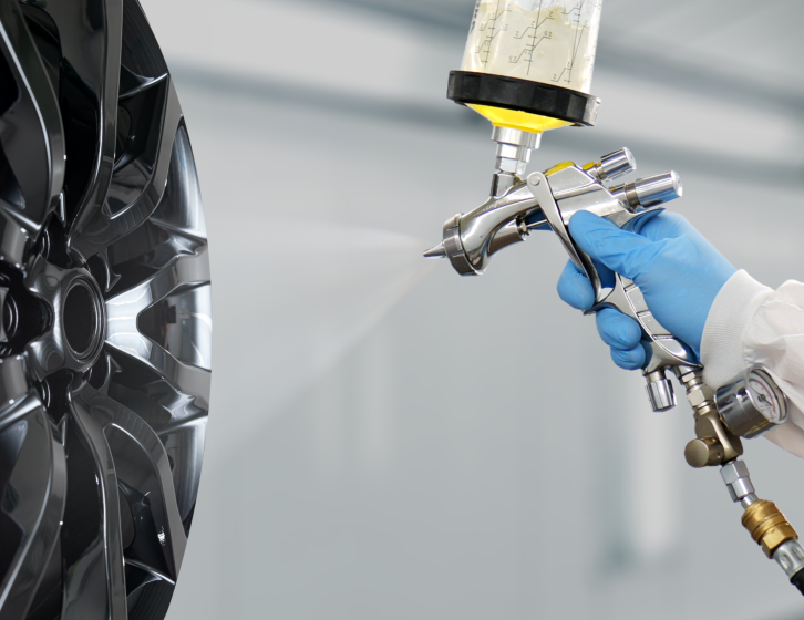 Gloved technician spraying a dark alloy wheel in a paint booth